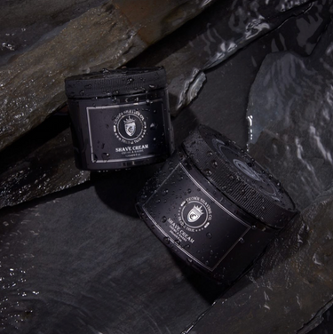 This image features two black jars of Crown Shaving Co. Shave Cream, partially submerged in water amongst dark, textured rocks. Water droplets cover the jars, and the labels with the brand's logo and product name are visible. The lighting creates a dramatic and moody atmosphere.